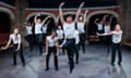 12 dancers in black trousers and white t-shirts, some jumping, arms raised, others knees bent, arms down