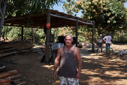 A man stands in a dusty open space with a shelter behind him over what appears to be a large pile of wood