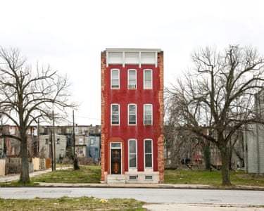 A tall, narrow red brick rowhouse in Baltimore stands alone, flanked by empty demolished lots that are grassed over with a trio of mature trees