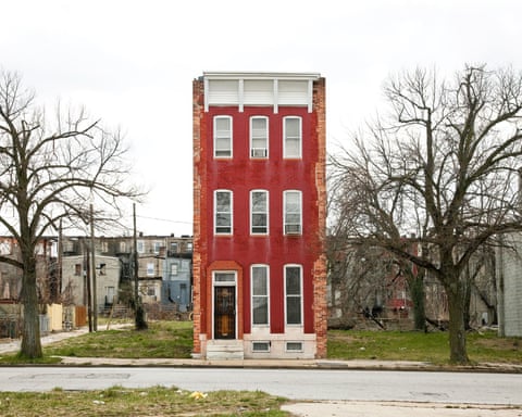 A tall, narrow red brick rowhouse in Baltimore stands alone, flanked by empty demolished lots