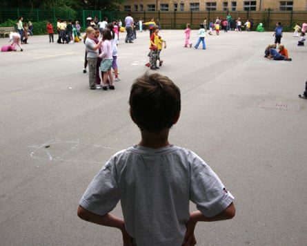 A child stands alone in a primary school playground as others play in the background. He is seen from behind in shadow.
