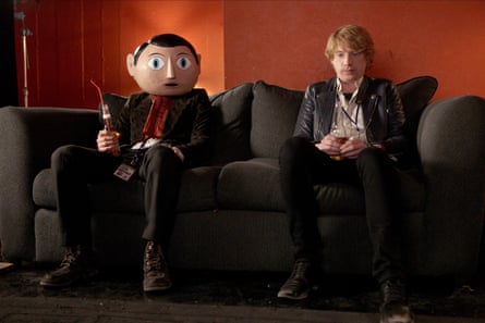 A man with a big cartoon head sitting beside a red-haired man on a dark sofa with an orange wall behind them