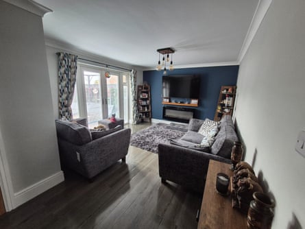 The refurbished living room: it is painted white with a teal-blue back wall, wooden flooring, grey sofa and armchair. There are wooden units with photos, vases and decorative objects, and patterned curtains. Everything looks brand-new.