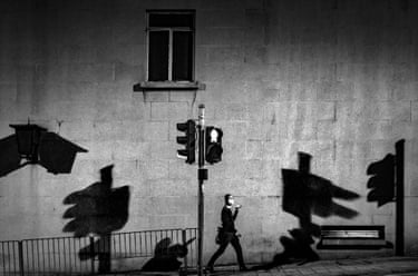 A man in a suit waers a facemask and talks on the phone while walking past shadows of traffic lights in a black and white image