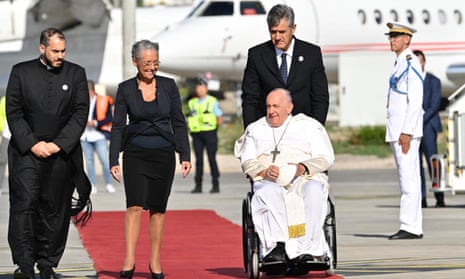 Pope Francis in a wheelchair on a red carpet next to Élisabeth Borne, with a plane in the background