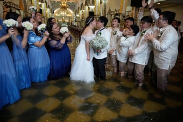 The newlyweds share a kiss as a line of bridesmaids in blue dresses and line of groomsmen in white suits either side of them cheer at their wedding in a floodied church