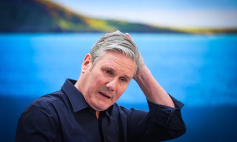 Keir Starmer in shirtsleeves, with one hand on the back of his head, speaking, against a backdrop of large photo of a lake