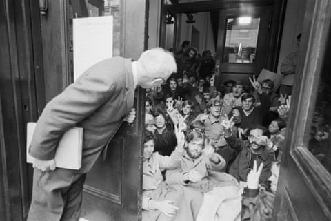 group of young people seated on hallway floor give peace sign as an older person, in the foreground, peers out at them from behind a door