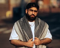 Rapper Fat Papi, bearded and in glasses, wears a patterned scarf over a white shirt outdoors