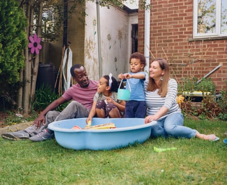 David and Rebecca Kitenda sit on the lawn in their garden either side of their children who are playing in a blue plastic sandpit