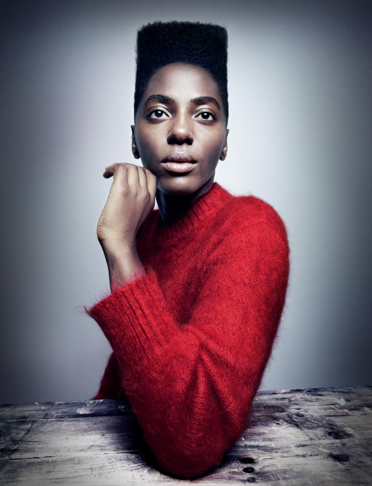 Ysra Daley-Ward  in a red jumper looking directly at the camera, her elbow on a table in front of her