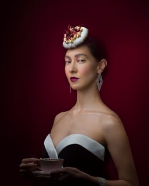 Female model with dark hair wearing a black and white cocktail dress and holding a pink tea cup and saucer. The model has a meringue with cream and poached pears dessert placed on her head like a hat