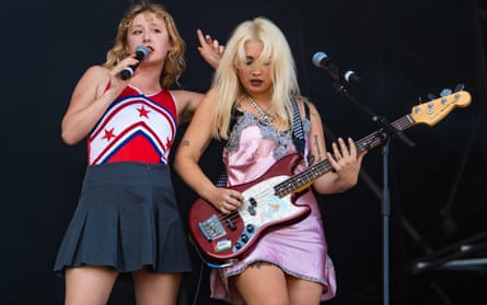 A young woman in a pink dress plays the bass guitar at a microphone while her bandmate, in a starry red-and-white top and a short dark skirt sings behind her