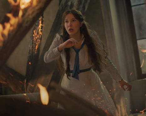 Movie still of a young woman in a white dress looking pained and surrounded by burning timbers.