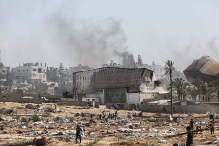 Smoke rises from bombed buildings as people wander around a desolate looking landscape