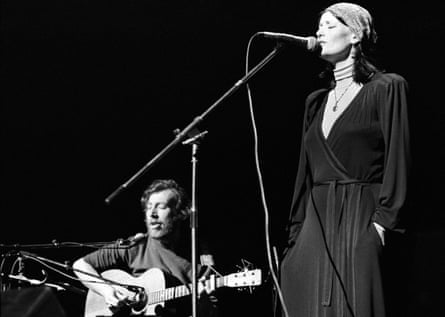 The Thompsons on stage, Richard sitting playing an acoustic guitar, Linda standing singing into a mic, eyes closed, wearing a long black dress and patterned headscarf.