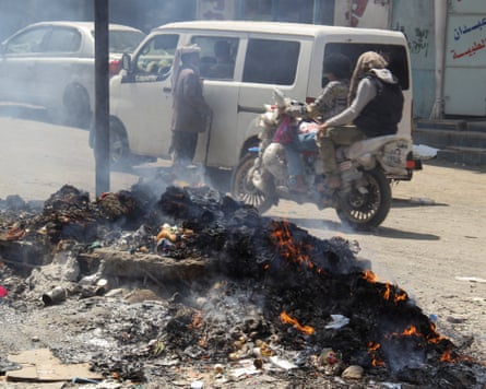 People on a motorbike ride past a pile of burning rubbish heaped up in a street