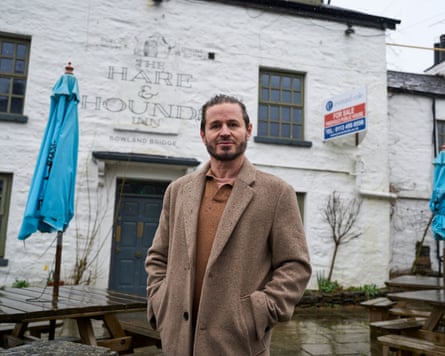 A man in a camel-coloured coat stands outside a pub with a for sale sign at the front