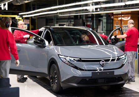 Four Nissan workers dressed in red tops inspect a shiny grey car.