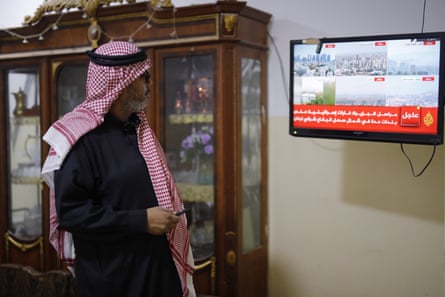 A middle-aged Arab man in a black thobe and red and white keffiyeh watches news of Gaza on the TV