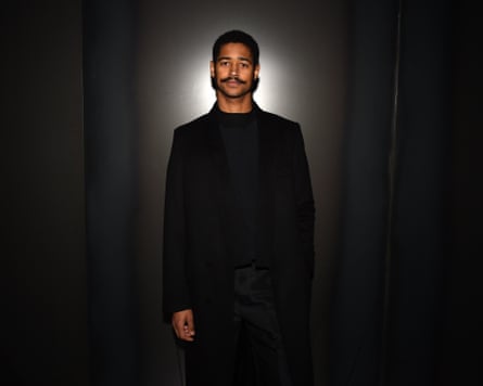 Alfreid Enoch wearing black and standing against a dark background looking at the camera.