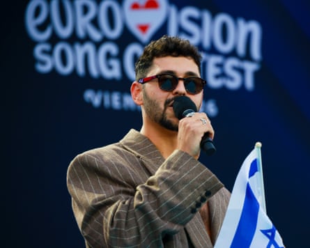 Noam Bettan sings wearing sunglasses and holding an Israeli flag against Eurovision song contest logo backdrop