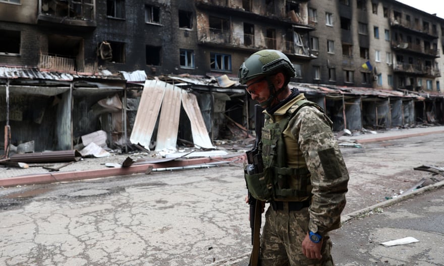 A Ukrainian soldier in uniform walks past a row of destroyed buildings