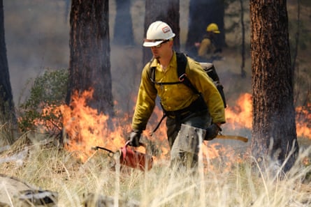A firefighter holding a red blowtorch walks through a field, fire in the background.