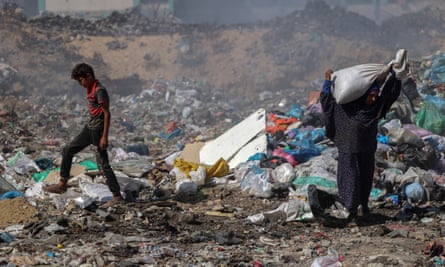Two people walk through a huge rubbish dump, with one carrying a large bag on their shoulder