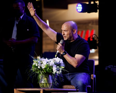 A bald man in a black T-shirt sits by a table with a vase with white flowers holding a microphone and his right hand raised in the air