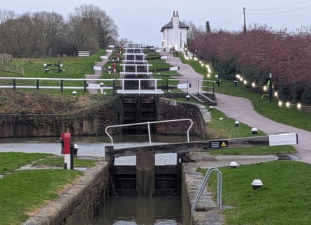 A series of locks on a canal, with grassy banks and a path lit by fairy lights going up the hill to a small white house