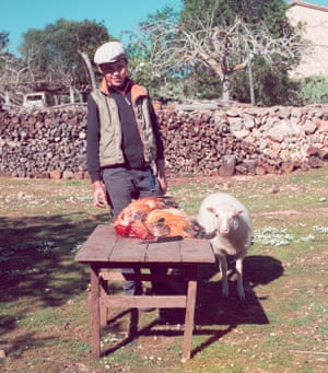 Miguel, in a flat cap and a down vest, a sheep at his side, in front of a table with a dead bird on it