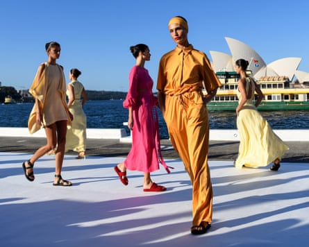 Models wearing summer clothes walk on a catwalk near the water with Sydney Opera house in the background