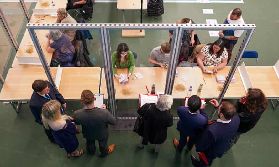 Cashiers are seated behind a clear screen. Seven people on the other side of the screen, some holding clipboards.