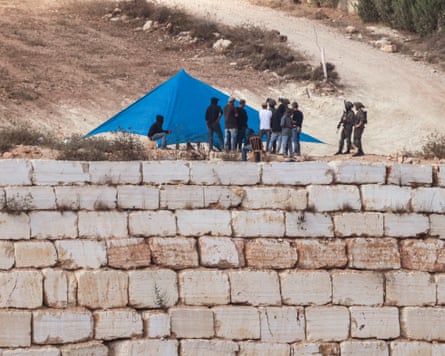 Israeli soldiers watch Israeli settlers setting up a tent just outside the entrance of a besieged Palestinian family home in the village of Qusra