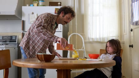 A man with a moustache pours orange juice into a glass for a bored looking child sitting at a table.