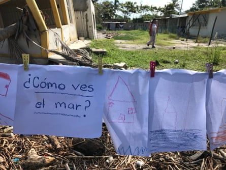 Children’s drawings of houses hang on a line, attached with pegs, with damaged buildings in the background. Alongside the drawings are the words, in Spanish, “How do you see the sea?”