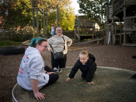 A laughing young woman in a hoodie on a roundabout with a small child as a teenaged girl in a hoodie looks on