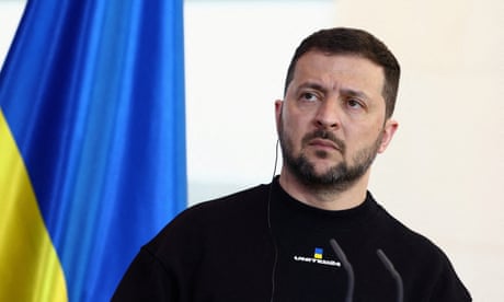 Ukraine's President Zelenskiy standing in front of a Ukraine flag with an earpiece in.