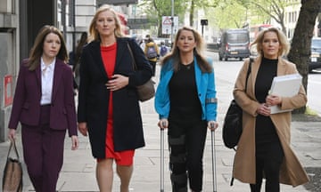 Four smartly-dressed female BBC news presenters walk side by side in a London street as they arrive for their tribunal case; McVeigh has long brown hair and wears a maroon trouser suit with white shirt; Croxall has long blond hair and wears a bright red dress under a black coat; Giannone is also blond and wears a sky-blue jacket over black top and trousers, and she is walking on crutches with a brace on one of her legs; Madera has shorter blond hair and wears a camel coat over black, and is carrying a large black handbag and thick white file of paperwork.
