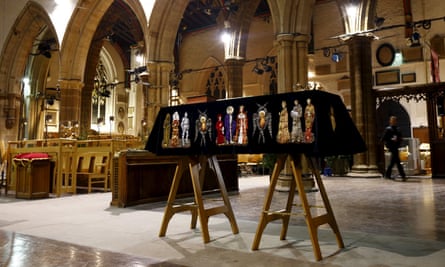 A coffin on two trestles with a crown on top of it, draped with an ornate black shroud with embroidered historical figures all the way around it, inside a cathedral