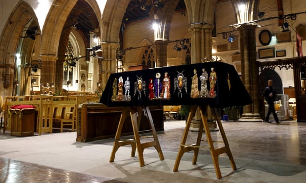A coffin on two trestles topped with a crown, draped in a black shroud decorated with embroidered historical figures all around, inside a cathedral