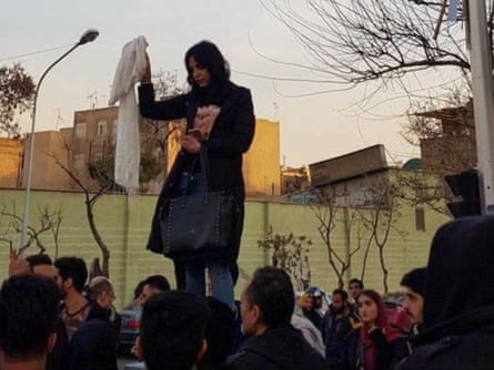 A woman with her head uncovered stands on a bin or postbox with passersby at her feet, holdinga hijab inthe air.