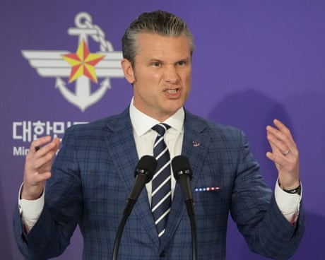 A grimacing man wearing a small suit gestures as he speaks into a microphone.