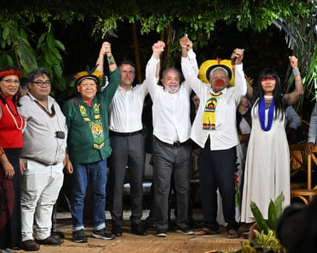 Lula and Macron hold their hands up with Indigenous leaders in a forest.
