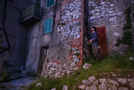 A man stands next to a stone house in the dark with his hand on the chimney breast.