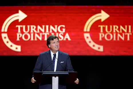 man in suit stands at lectern, a red screen saying 'turning point usa' behind him