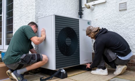 Two technicians installing a heat pump unit at a 1930s-built house in Folkestone, UK; they are crouching down and positioning it in front of a white wall