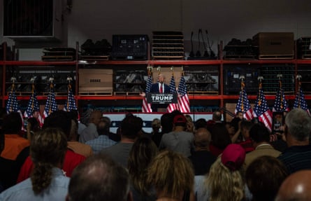 Donald Trump, in a blue suit with a red tie, stands surrounded by American flags before a crowd of people in Clinton, Michigan.