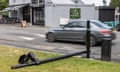 A car drives past a Ulez camera on a post that is damaged and lying on the grass with a Smoky Boys Bar and Grill across the street